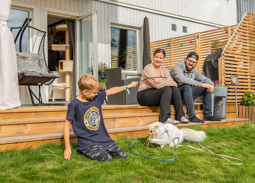 Porträtt på en familj som sitter på sitt trädäck och sonen leker med katten i gräsmattan