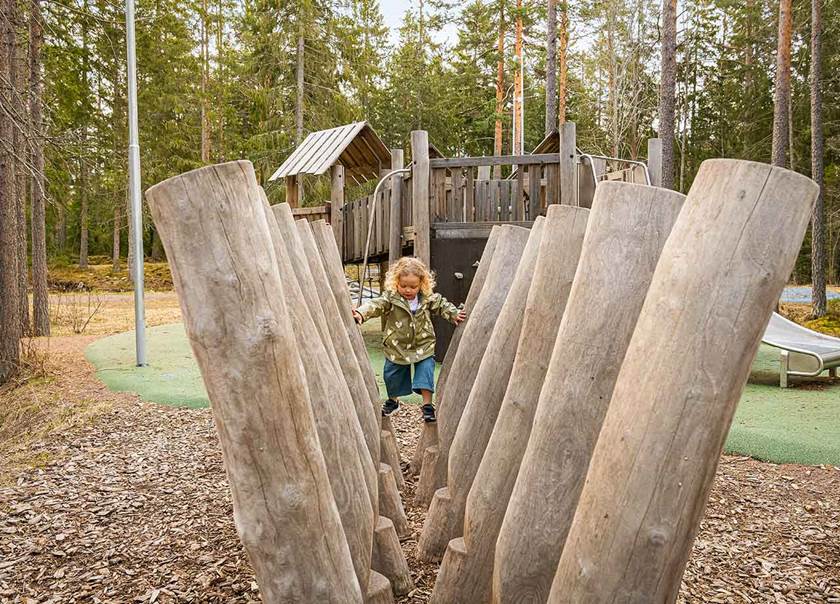 Liten flicka i grön regnjacka och blont hår går balansgång på stora stockar.