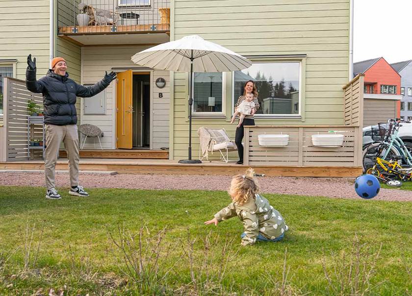 Pappa i svart jacka och beige byxor spelar fotboll leker med flicka i grön regnjacka och blont hår.