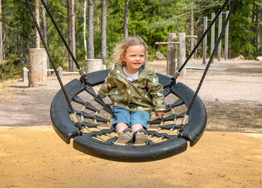 Liten flicka i blont hår och grön regnjacka och jeans gungar i stor gunga med nät.