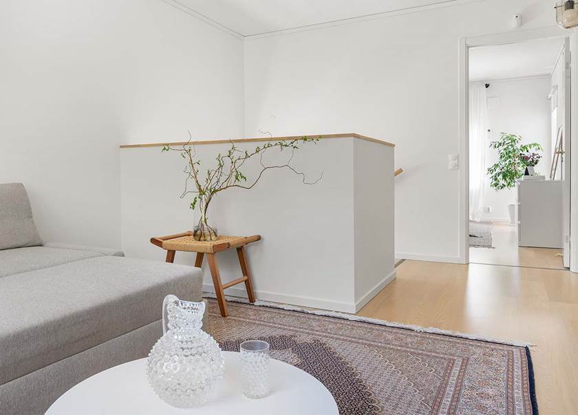 Ett allrum på en ovavåning är möblerad med en grå soffa på en orientalisk matta i grått, svart, rött och vitt. På ett sidobord står en glasvas med en trädgren i.