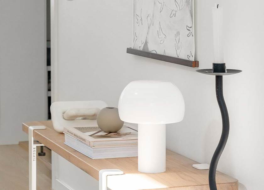 Smalt naturfärgat hallbord med en glaslampa i vitt och böcker. På golvet bredvid står en lång svart ljusstake.