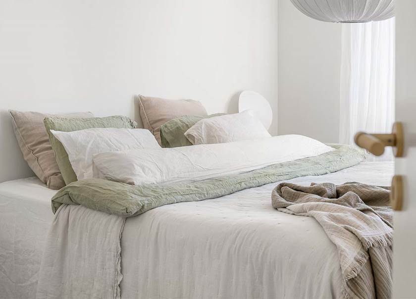 På en dubbelsäng med vita sängkläder ligger en filt i ljust brun färg. I taket hänger en klotformad lampa i vitt.